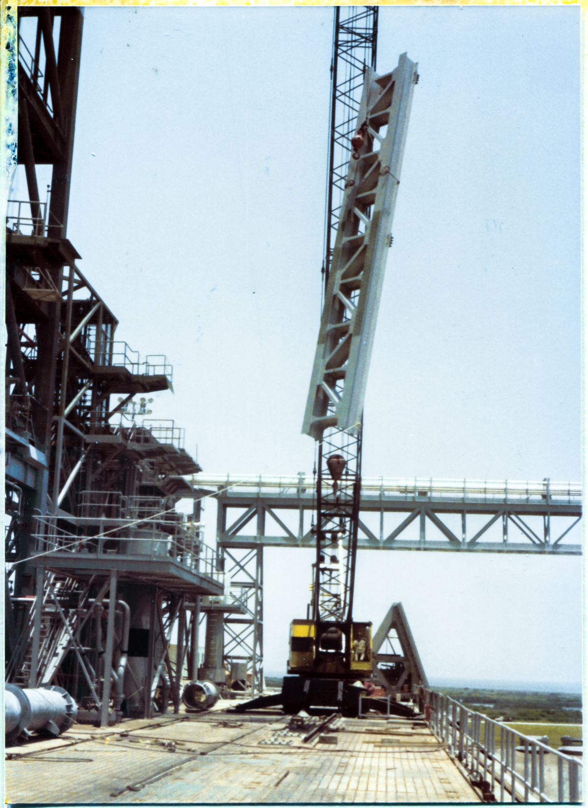 Image 078. The GOX Arm Strongback now begins its vertical journey to the top of the Fixed Service Structure (a small part of which is visible along the left margin of the photograph, extending up to and beyond the top) at Space Shuttle Launch Complex 39-B, Kennedy Space Center, Florida. Beneath it, on the ground, all personnel have been cleared from the area, and will remain so as a safety precaution until the Strongback is firmly welded to the FSS Primary Framing, far above where it is located at this moment. A Union Ironworker from Local 808, working for Ivey Steel, remains unseen, handling the tag line which is visible, attached to the lower right-side corner of the Strongback, keeping it properly oriented as it rises higher and higher into the sky. Photo by James MacLaren.